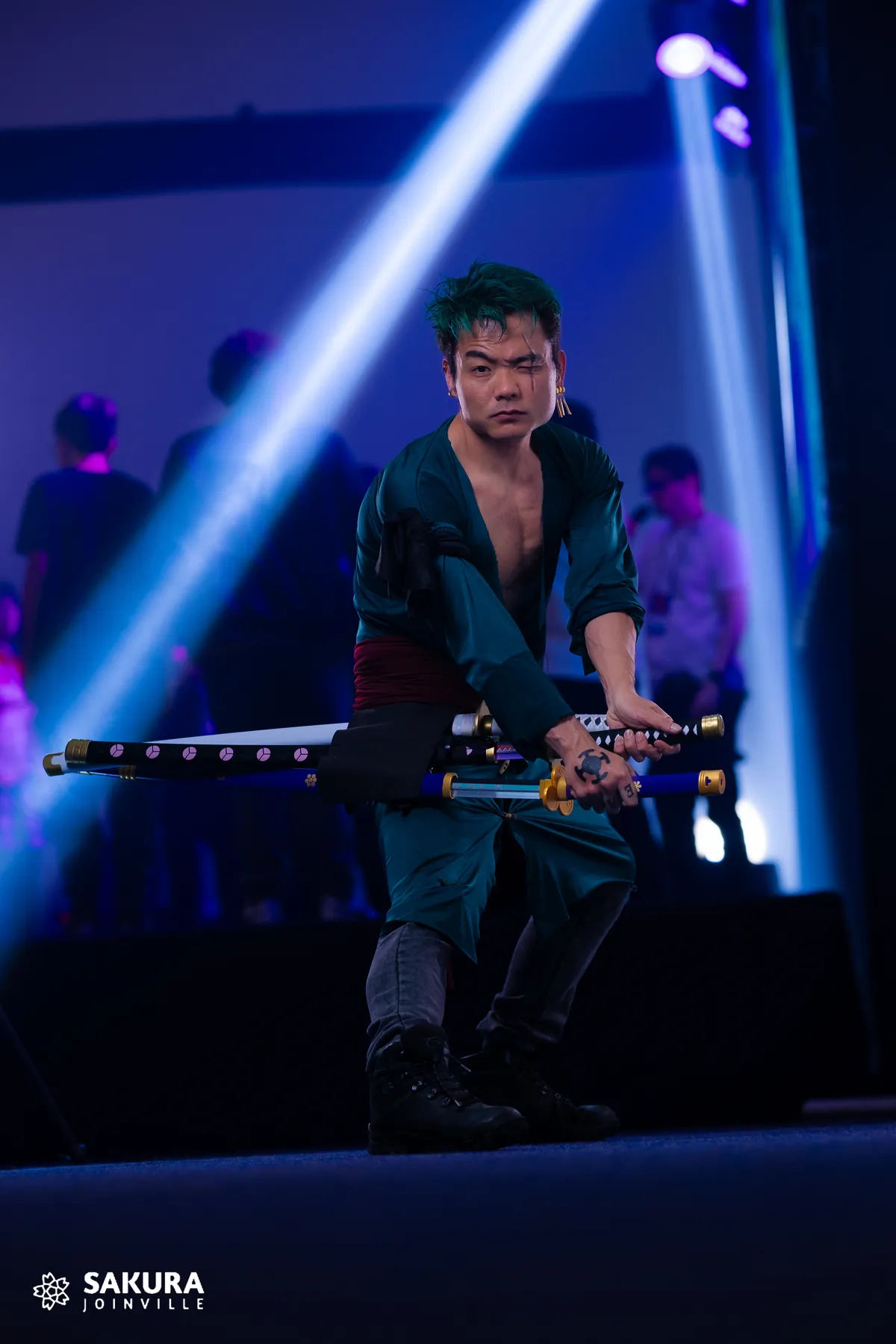 Eduardo em cosplay com espadas sobre um palco iluminado em azul